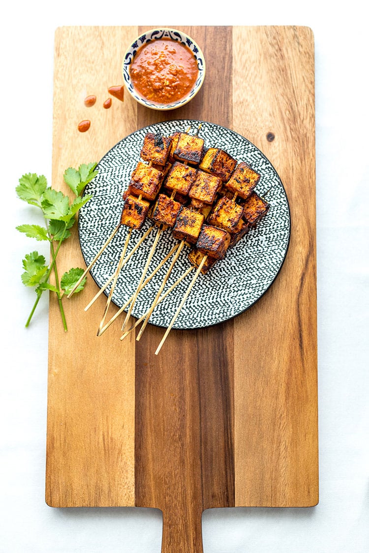 A pile of orange chipotle tofu skewers sit on top of a wooden chopping board on a white background. 