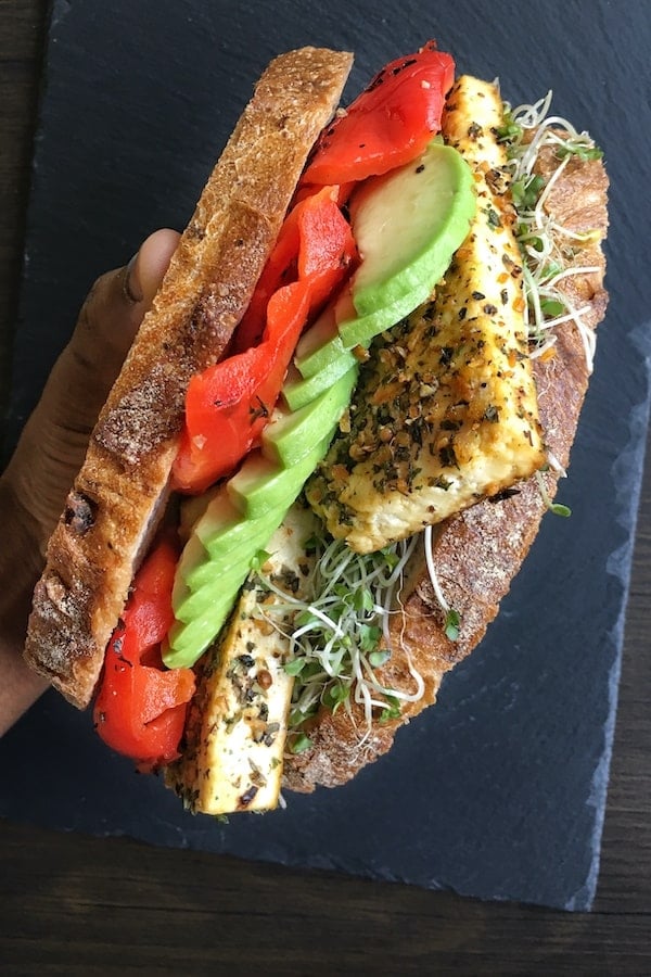 Crusty bread slices stacked between tofu, avocado and roasted red peppers held together with a hand. 