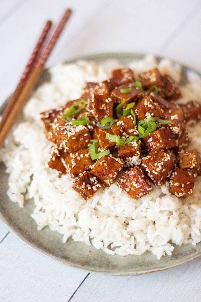 Garic tofu with seseme seeds sits on top of a plate of white rice. Topped with finely sliced spring onions the chopsticks sit on the side.