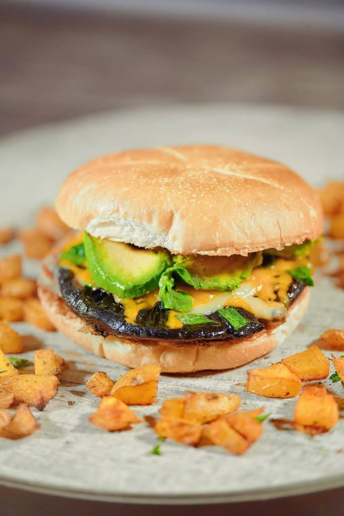 A vegan portobello mushroom burger sat on a textured wooden surface surrounded by sweet potatoes. 