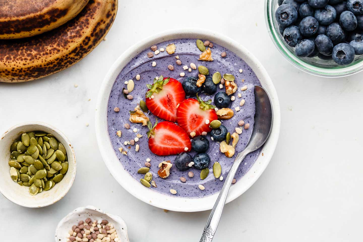 A bowl of vegan yogurt purple in colour topped with nuts, seeds, strawberries and blueberries which also surround the bowl.