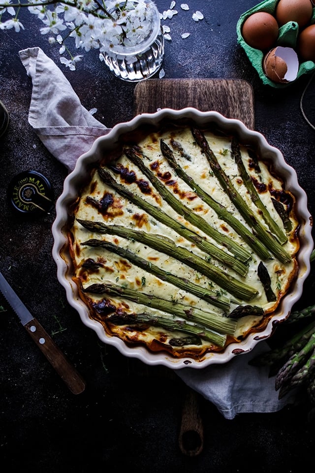 An entire Asparagus Quiche with Sweet Potato Crust sits inside a pie dish on a chopping board with a dark black background. At the side there's some cracked eggs, a decorative flower and a napkin.