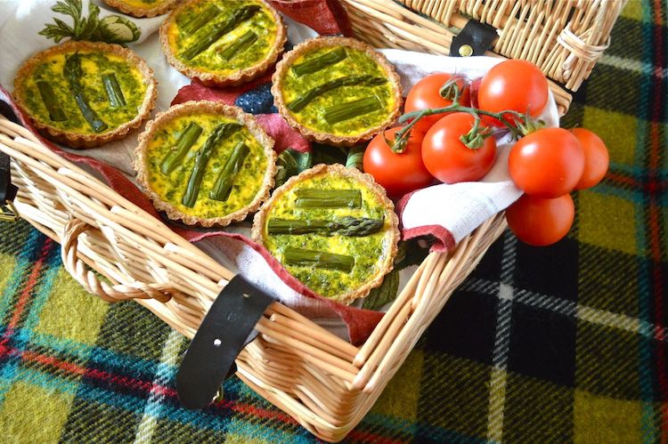 Five mini  Asparagus Tarts With A Pesto Surprise sit inside a wicker basket. At the side are some on the vine cherry tomatoes. The basket sits on a deocrative checked rug. 