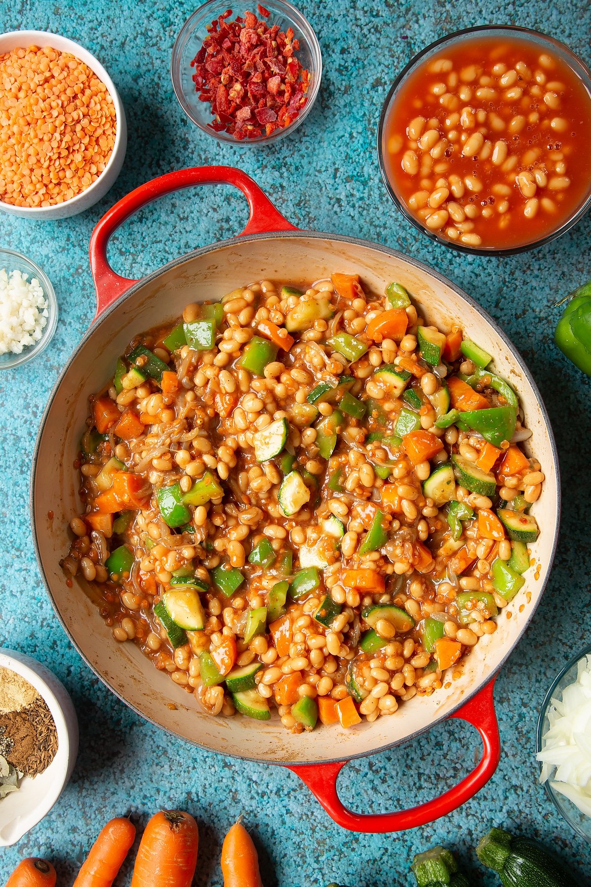 Fried onions, spices, peppers, carrots, courgette, red lentils and baked beans in a pan. Ingredients to make baked bean curry surround the pen.