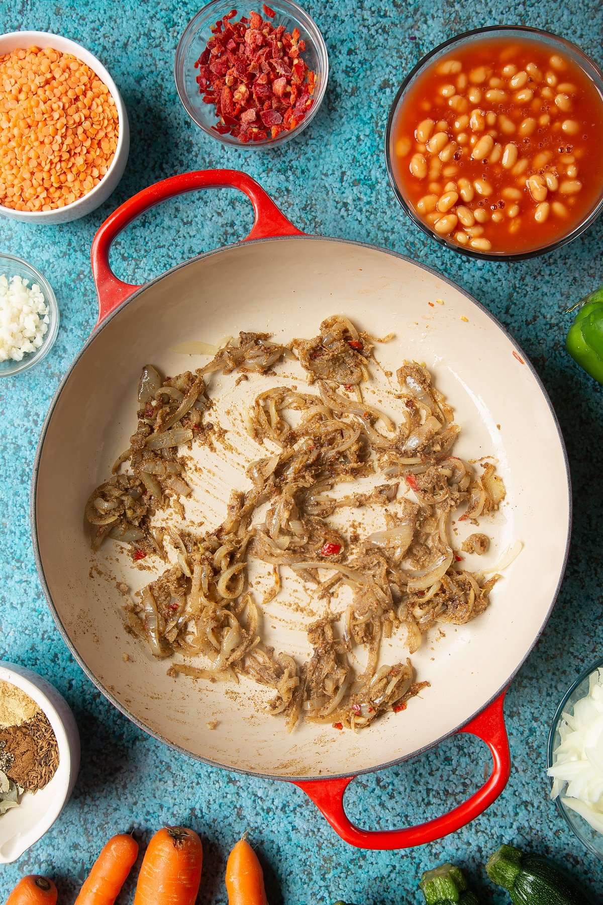 Fried onions, chilli, ginger, garlic and spices in a pan. Ingredients to make baked bean curry surround the pen.