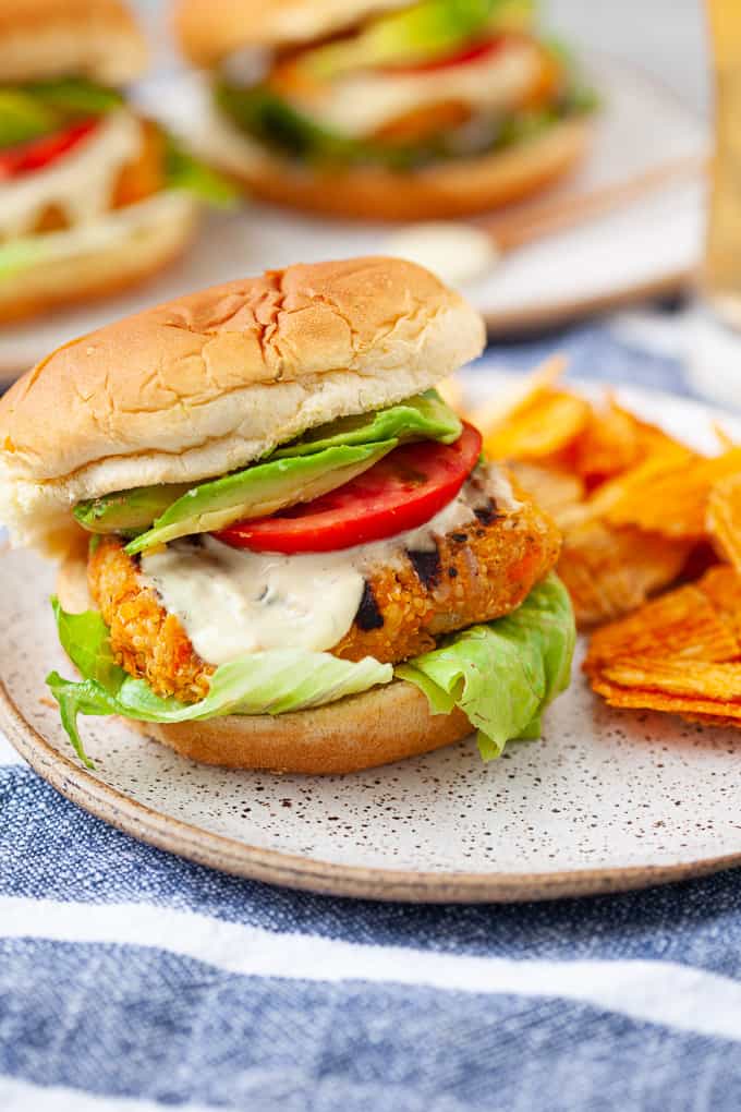 Buffalo chickpea burger on a decorative plate with a side of crisps. Two additional burgers are out of focus in the background.