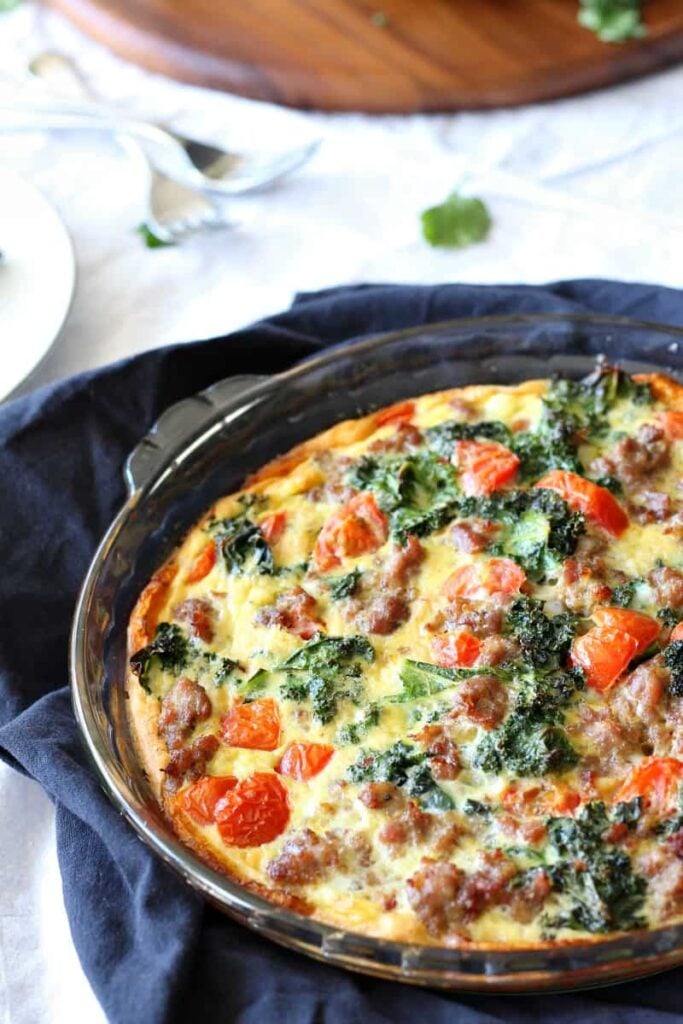 A whole butternut crust quiche with sausage, kale and tomatoes in a pyrex dish on a dark blue napkin. 