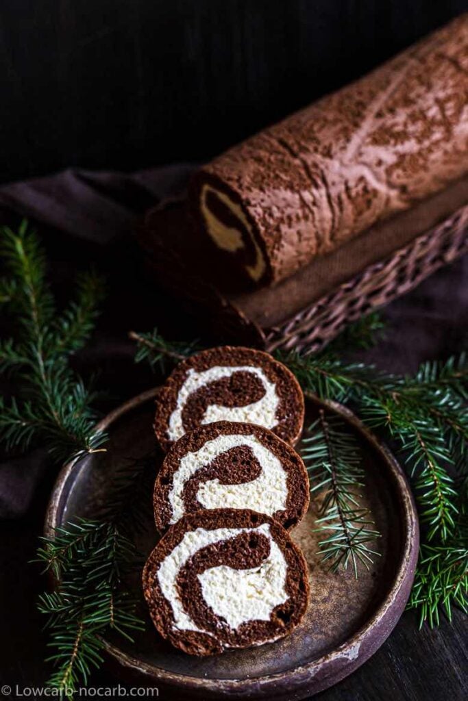 Three slices of keto chocolate Swiss roll sit ontop of a decorative dark plate. The side of the plate is deocrated with foliage. In the background you can see the whole Swiss roll which is sat ontop of a wicker serving board.