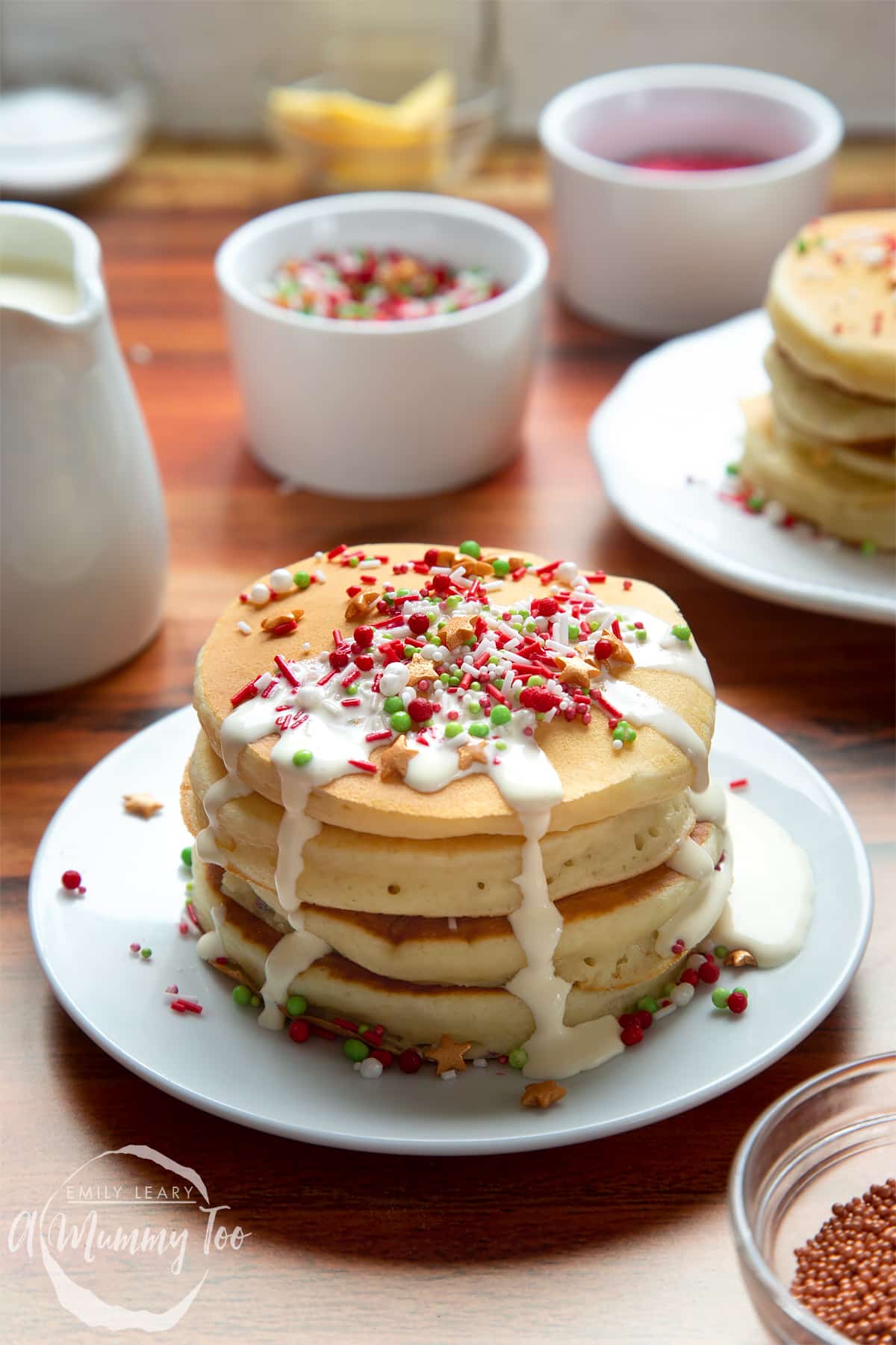 A tall stack of Christmas morning pancakes, stacked on a white plate, scattered with festive sprinkles and drizzled with cream.