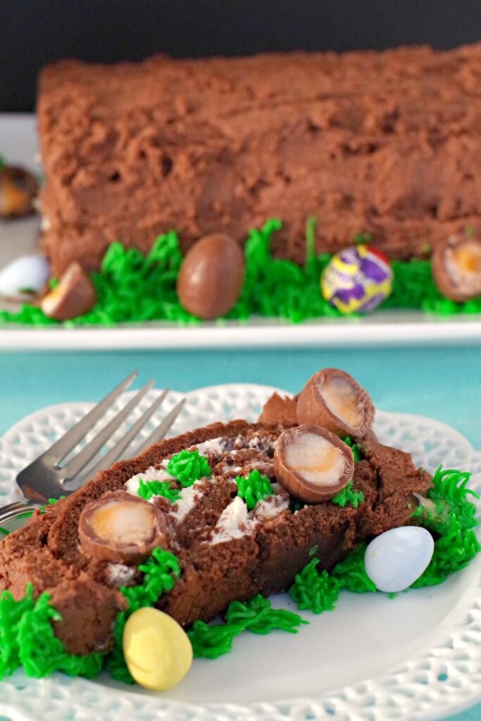 A slice of the easter egg chocolate roll sits on a white decorative plate covered in candy eggs and cadbury's cream eggs. In the background you can see the entire roll which sits on a white rectangular plate with a blue table cloth.