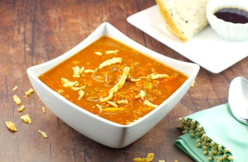 A square bowl of Creamy Carrot Soup on a dark brown wooden tabletop. At the side is a mint green napkin and a plate of bread with dip. 