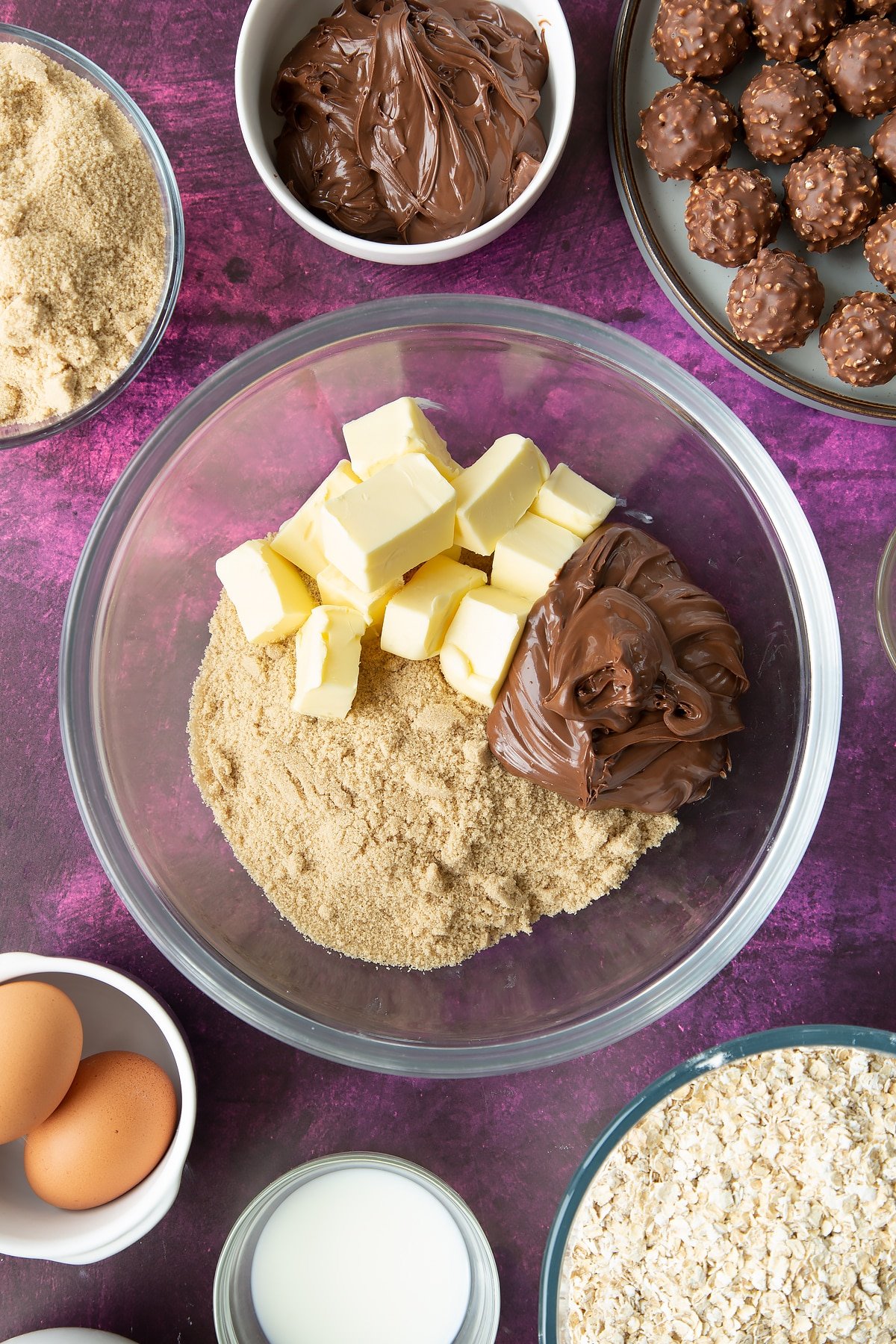 Soft brown sugar, Nutella and butter in a large mixing bowl. Ingredients to make Ferrero Rocher cookies surround the bowl.