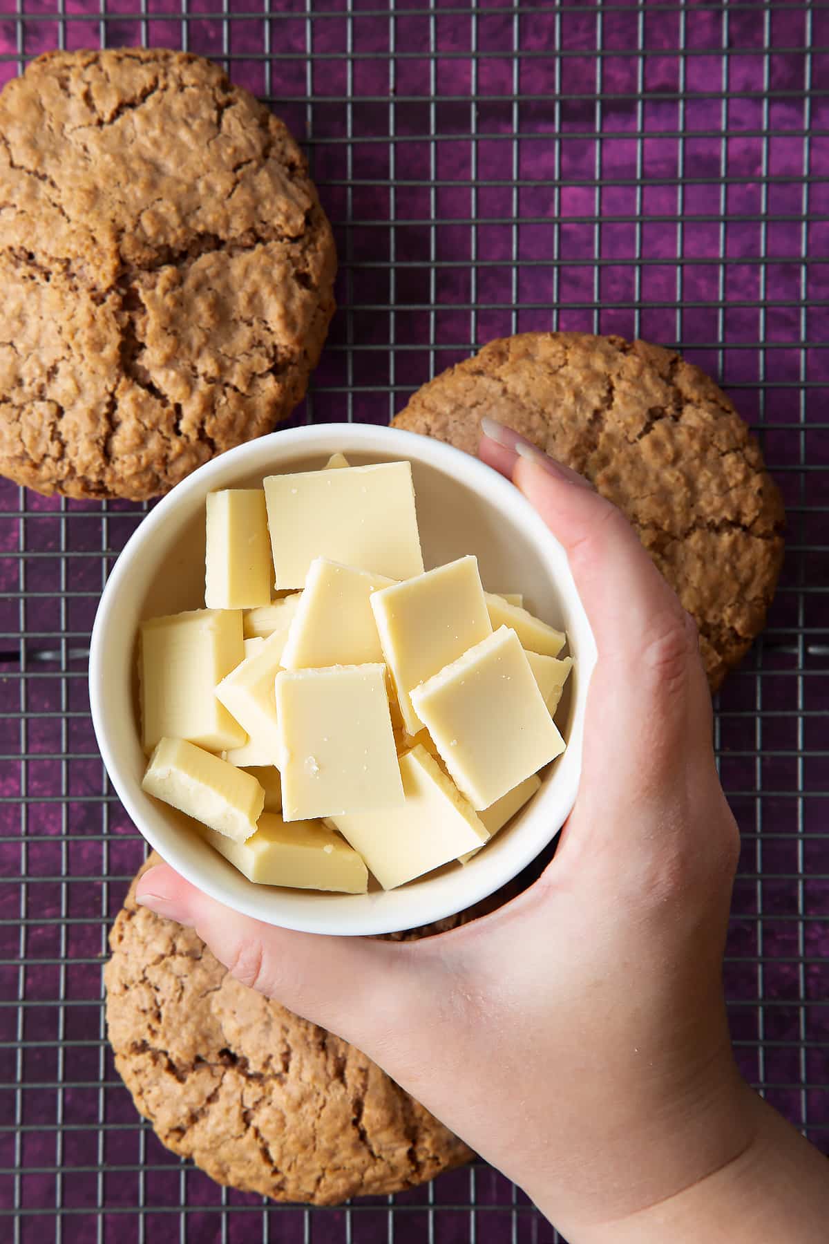 Hand holding a small bowl of white chocolate squares. Below, three undecorated Ferrero Rocher cookies sit on a cooling rack.