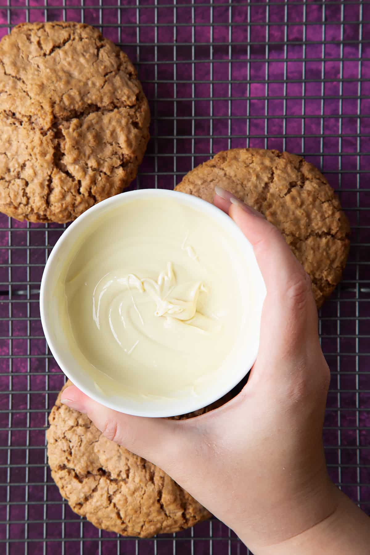 Hand holding a small bowl of melted white chocolate. Below, three undecorated Ferrero Rocher cookies sit on a cooling rack.