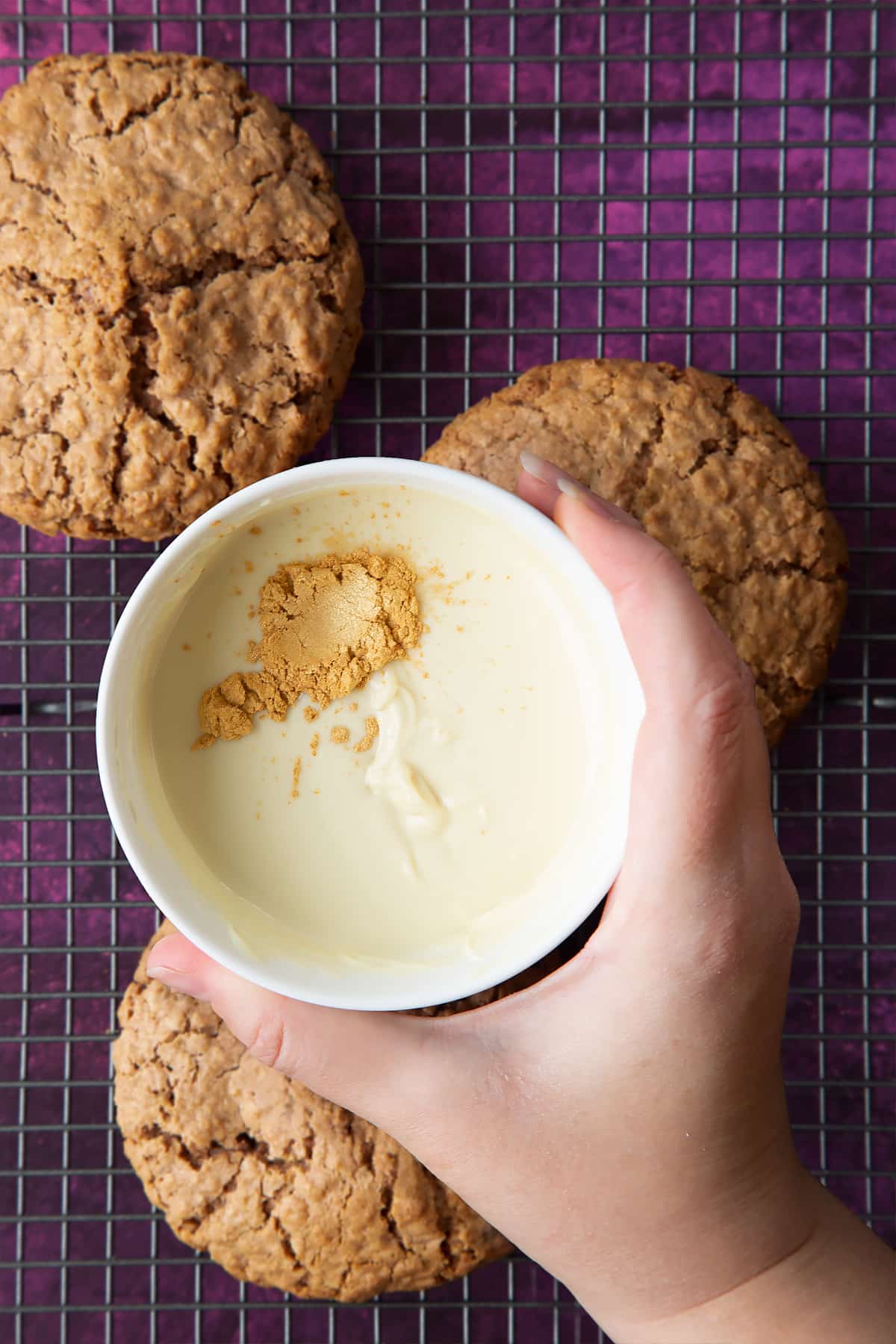 Hand holding a small bowl of melted white chocolate with gold lustre. Below, three undecorated Ferrero Rocher cookies sit on a cooling rack.