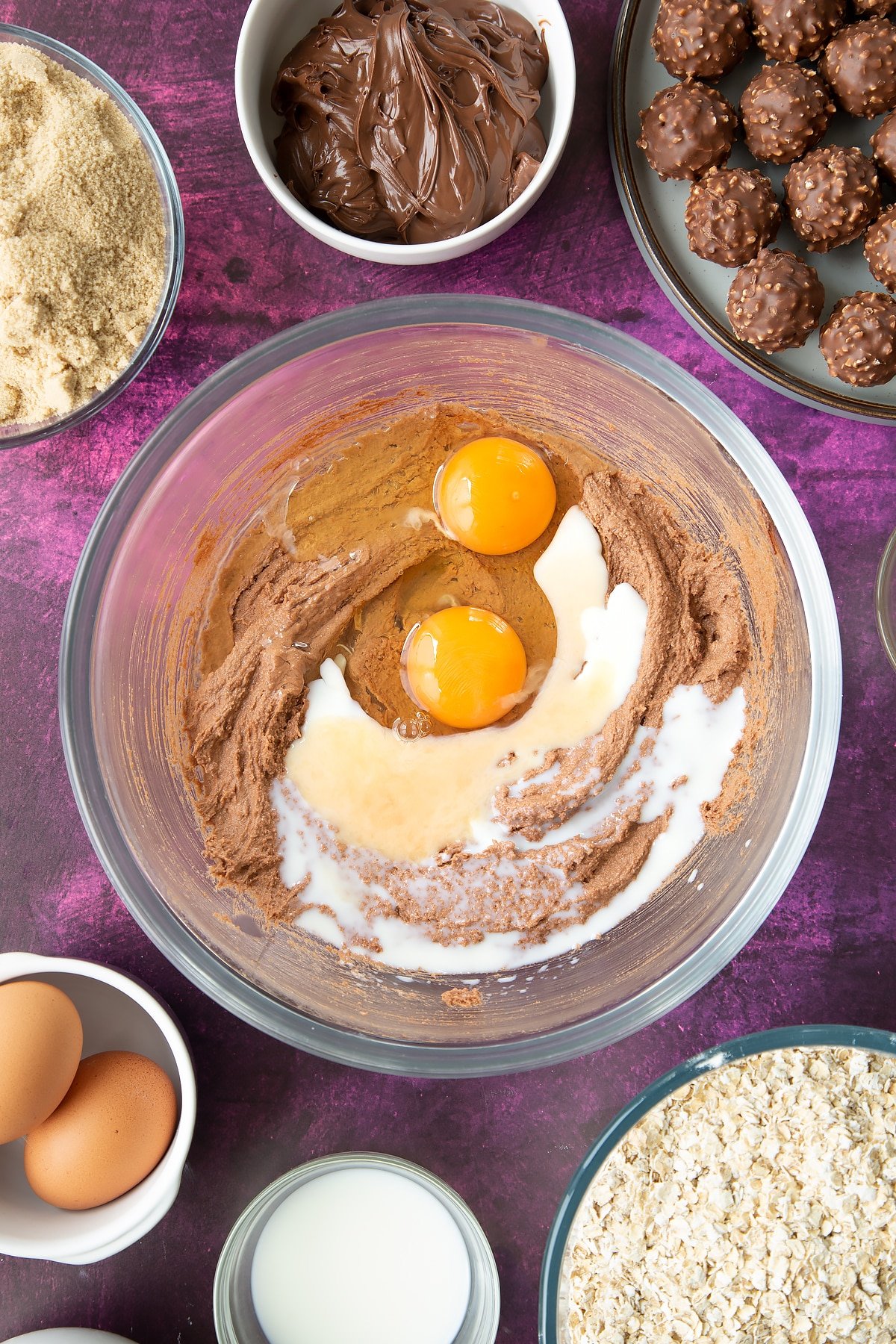 Soft brown sugar, Nutella and butter beaten together in a large mixing bowl with eggs and milk on top. Ingredients to make Ferrero Rocher cookies surround the bowl.