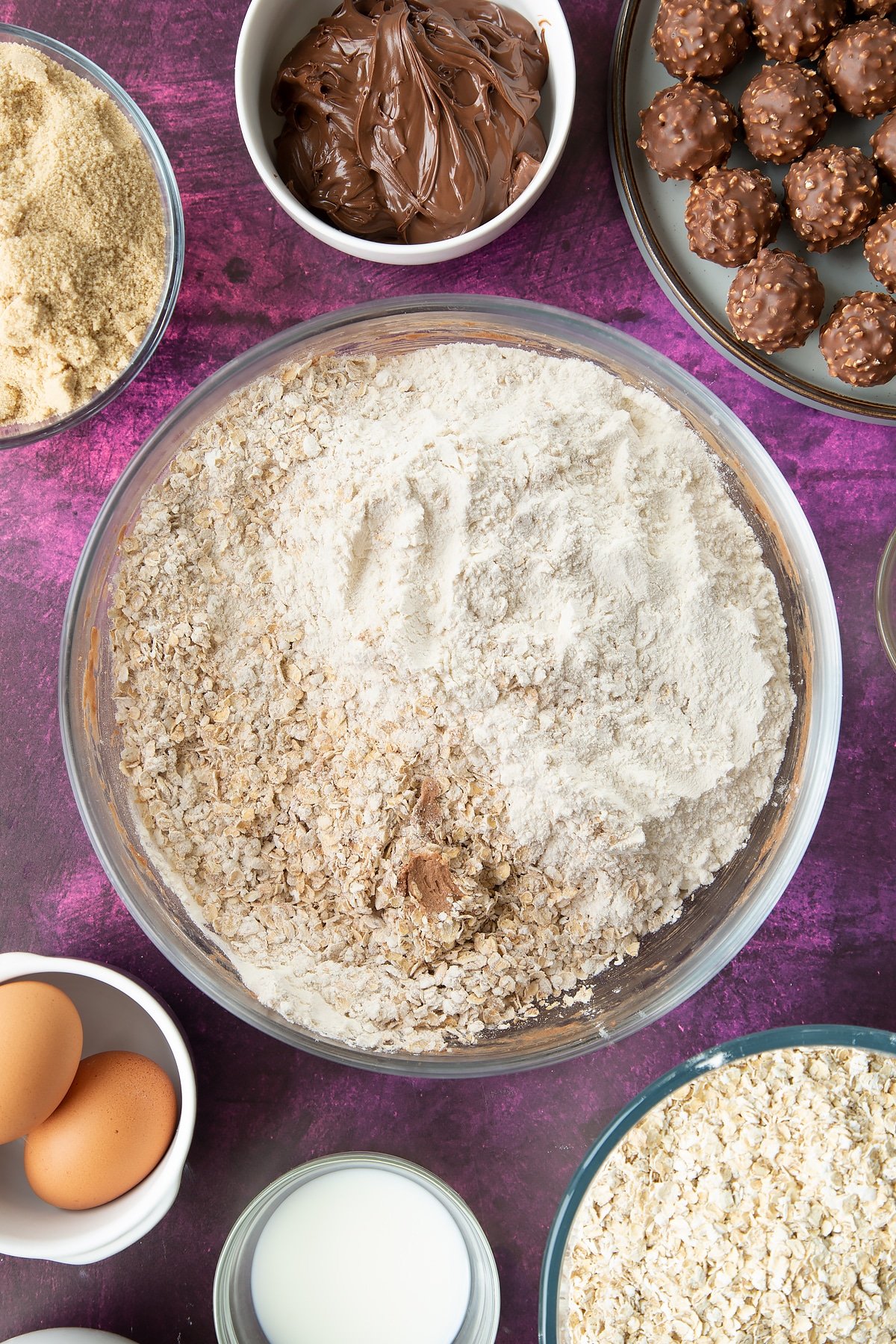 Soft brown sugar, Nutella, butter, eggs and milk beaten together in a large mixing bowl with flour and oats on top. Ingredients to make Ferrero Rocher cookies surround the bowl.