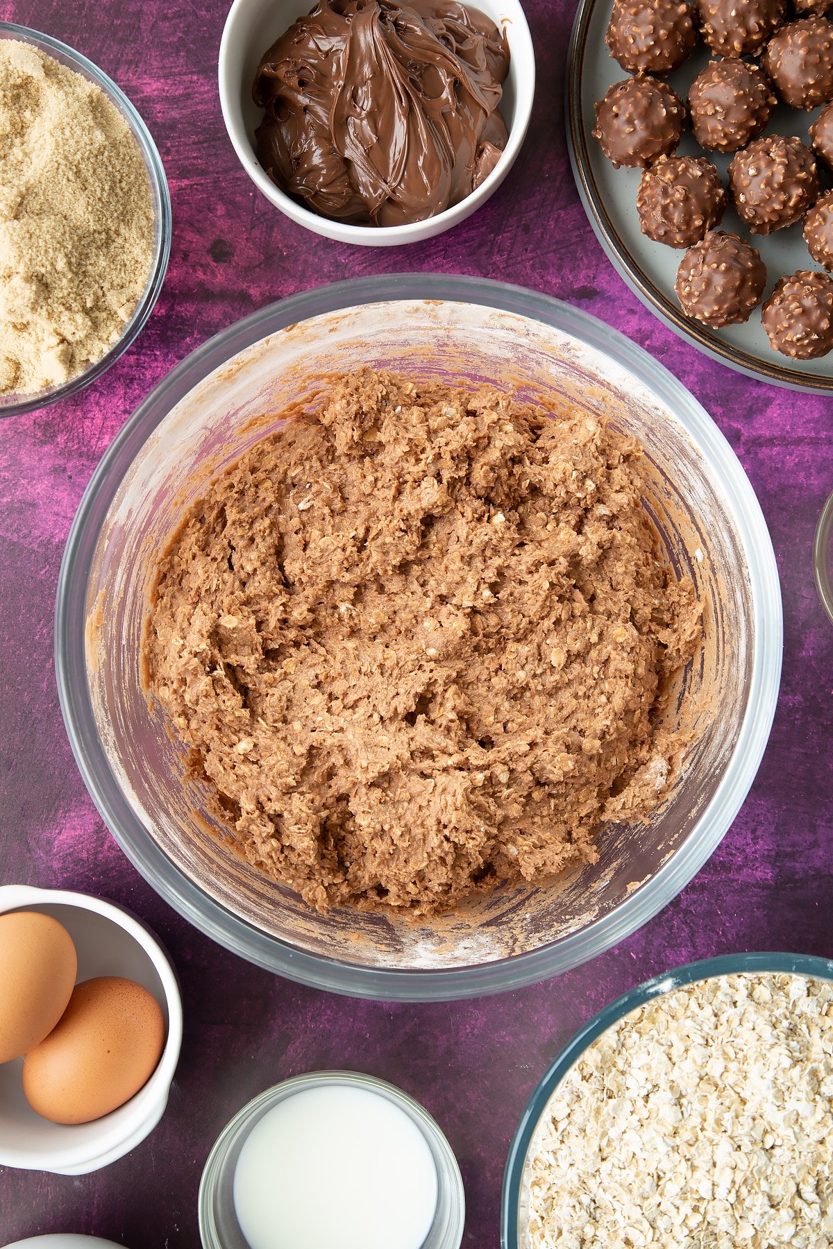 Oaty chocolate cookie dough a large mixing bowl with flour and oats on top. Ingredients to make Ferrero Rocher cookies surround the bowl.