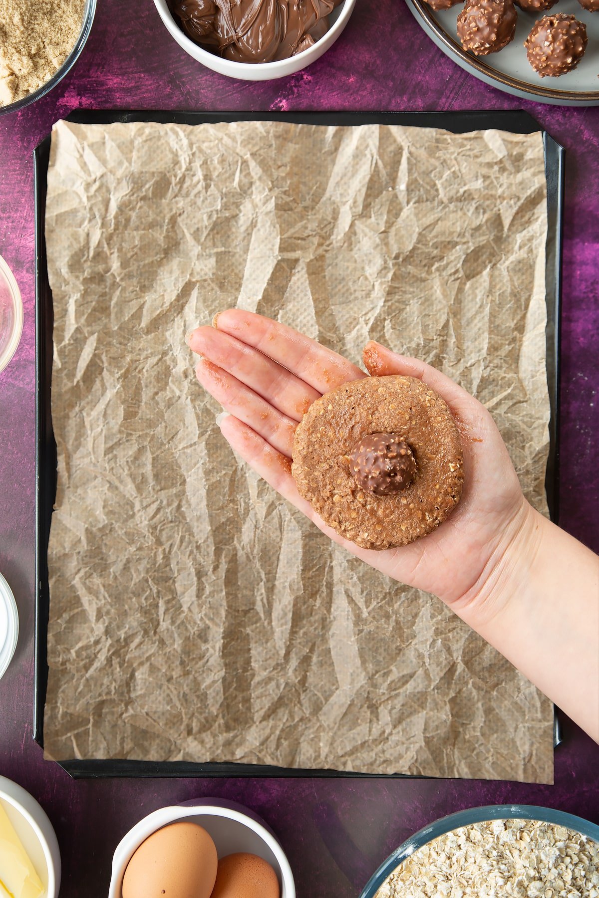 A hand holding a disc of oaty chocolate cookie dough with a Ferrero Rocher chocolate in the centre. Ingredients to make Ferrero Rocher cookies surround a tray below.