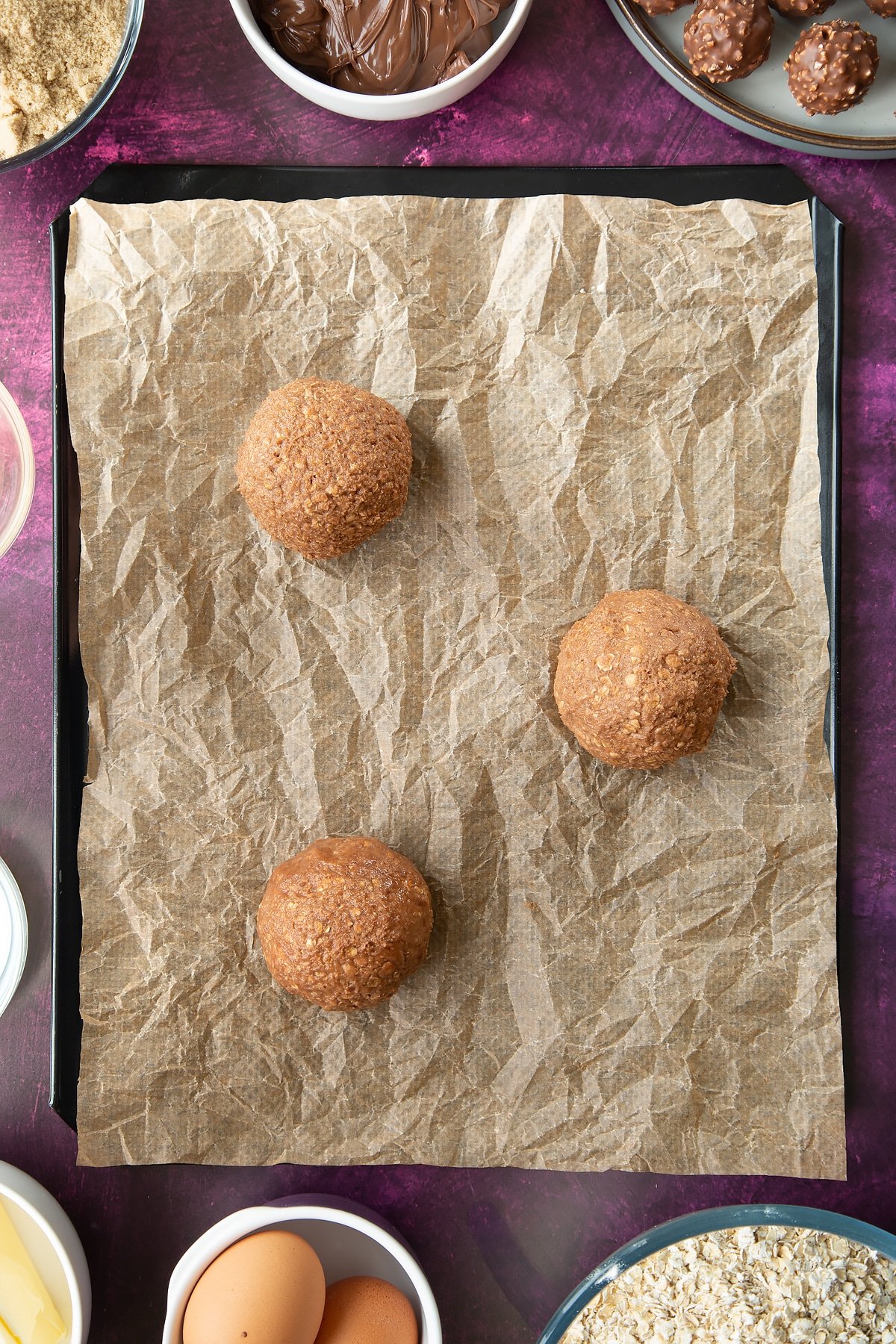 Three balls of Ferrero Rocher cookie dough on a lined tray. Ingredients to make Ferrero Rocher cookies surround the tray.