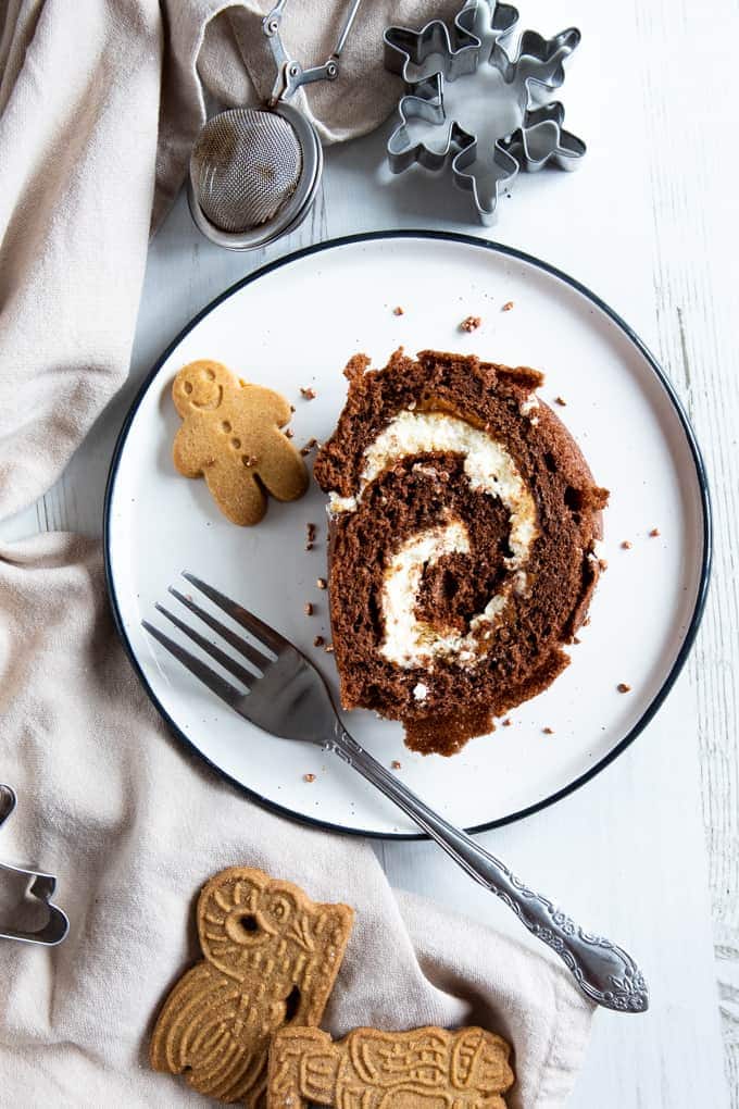 A slice of gingerbread chcolate log on a white plate. At the side is a minitiure gingerbread man and a decorative fork. The plate sits on a white wooden background with a table cloth at the side. At the top of the image there's a cute cookie cutter.