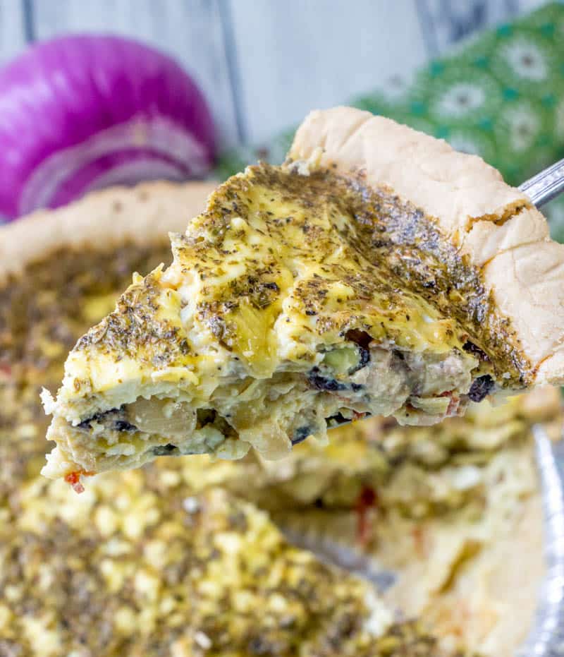 A slice of greek quiche being pulled out from the rest of teh quiche below using a large serving spoon. In the background there's a decorative napkin and a red onion.