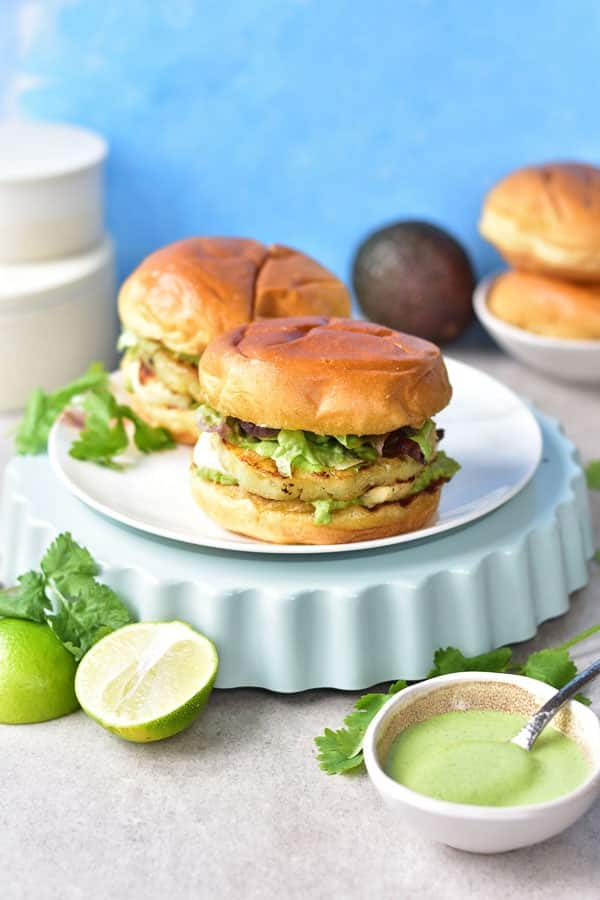 2 halloumi burgers on a white plate with limes and avocado dip in the foreground and a whole avocado in the background.