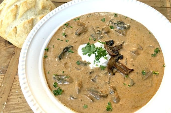 White bowl of Rustic Hungarian Mushroom Soup on a wooden surface with a bread roll on the side. 