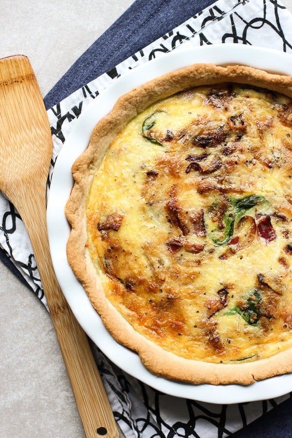 Bacon Spinach & Cheddar Quiche on a white plate. At the side is a wooden spatula. Under the plate are two napkins a blue one and a white and black one. 