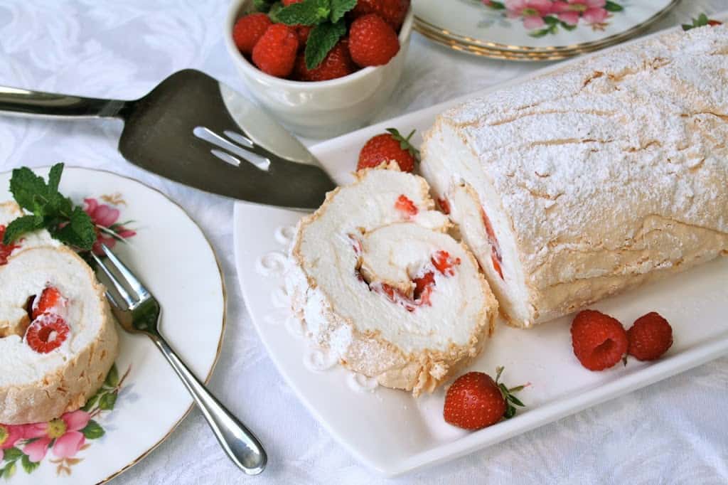 A summer berry meringue roulade sits on a large decorative white plate on top of a white table cloth. The side is decorated with raspberries and strawberries. At the side there's a floral decorative plate with a slice of the Swiss roll and a cake fork.