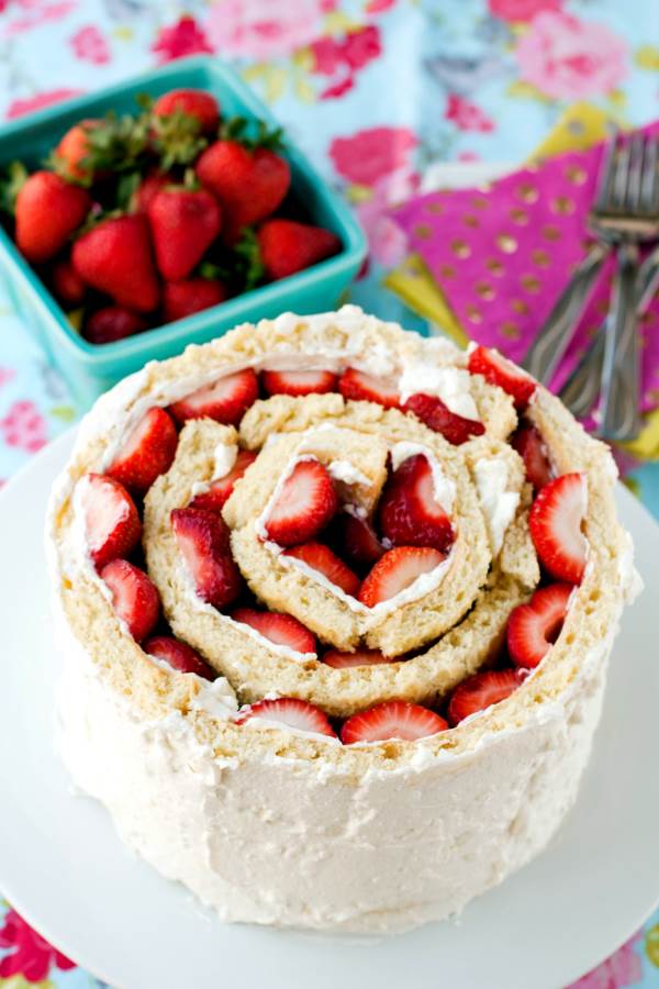 A strawberry shortcake roll-up cake sits on a white round plate. At the side there's a bowl of strawberries and some forks sitting ontop of some colourful decorative napkins.