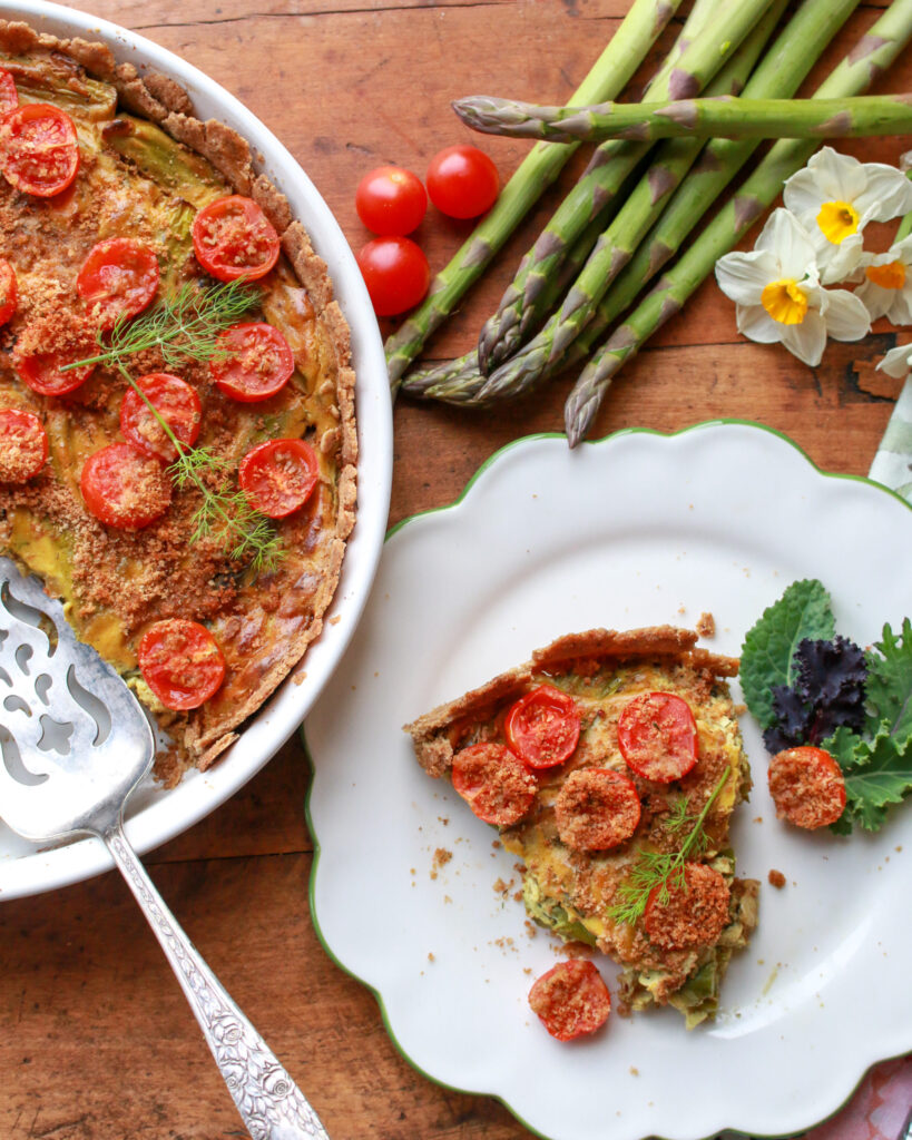 One slice of Asparagus Dill Tofu Quiche on a decorative white plate with a side salad. There's a large dish of Asparagus Dill Tofu Quiche on the otherside of the image. At the top there's some asparagus. 