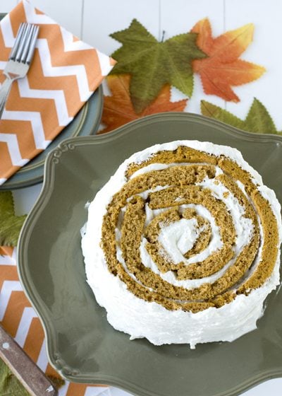 A pumpkin slice on a olive green deocrative plate on a white wooden table. The table is deocrated with orange and white napkins and green and orange leaves.