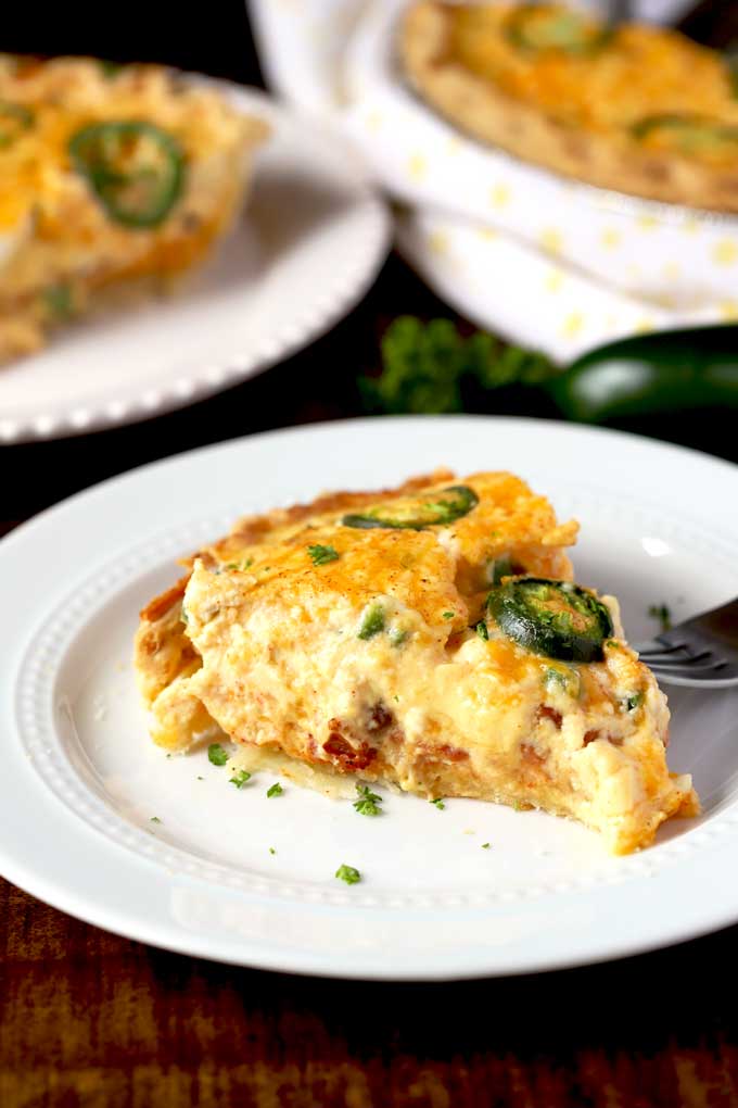 A slice of Jalapeno Popper Quiche sits on a white plate with a fork at the side in the foreground. You can see two additional Jalapeno Popper Quiche slices on plates in the background which are out of focus.