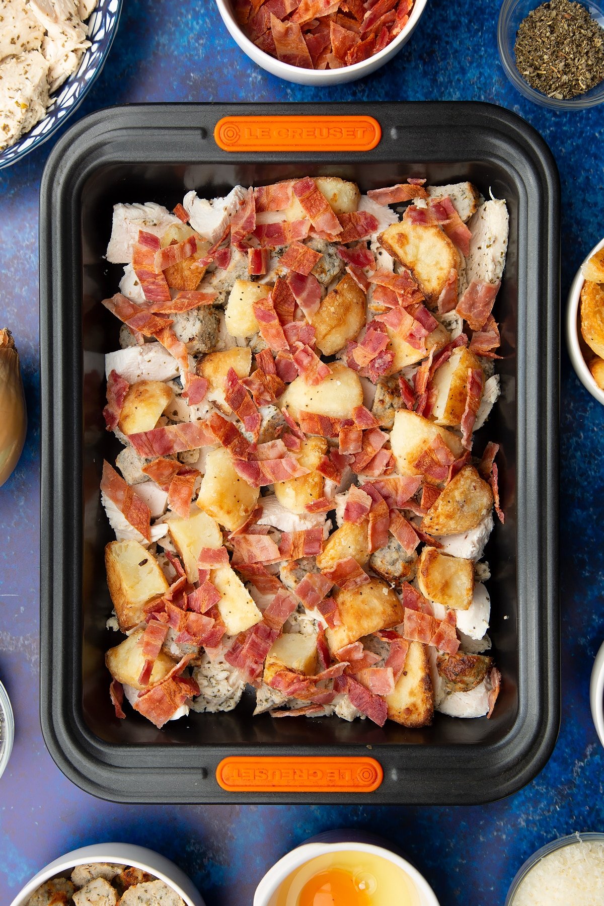 Chopped cooked turkey, stuffing, bacon and roast potatoes in a roasting dish. Ingredients to make a leftover Christmas dinner pie surround the tray.