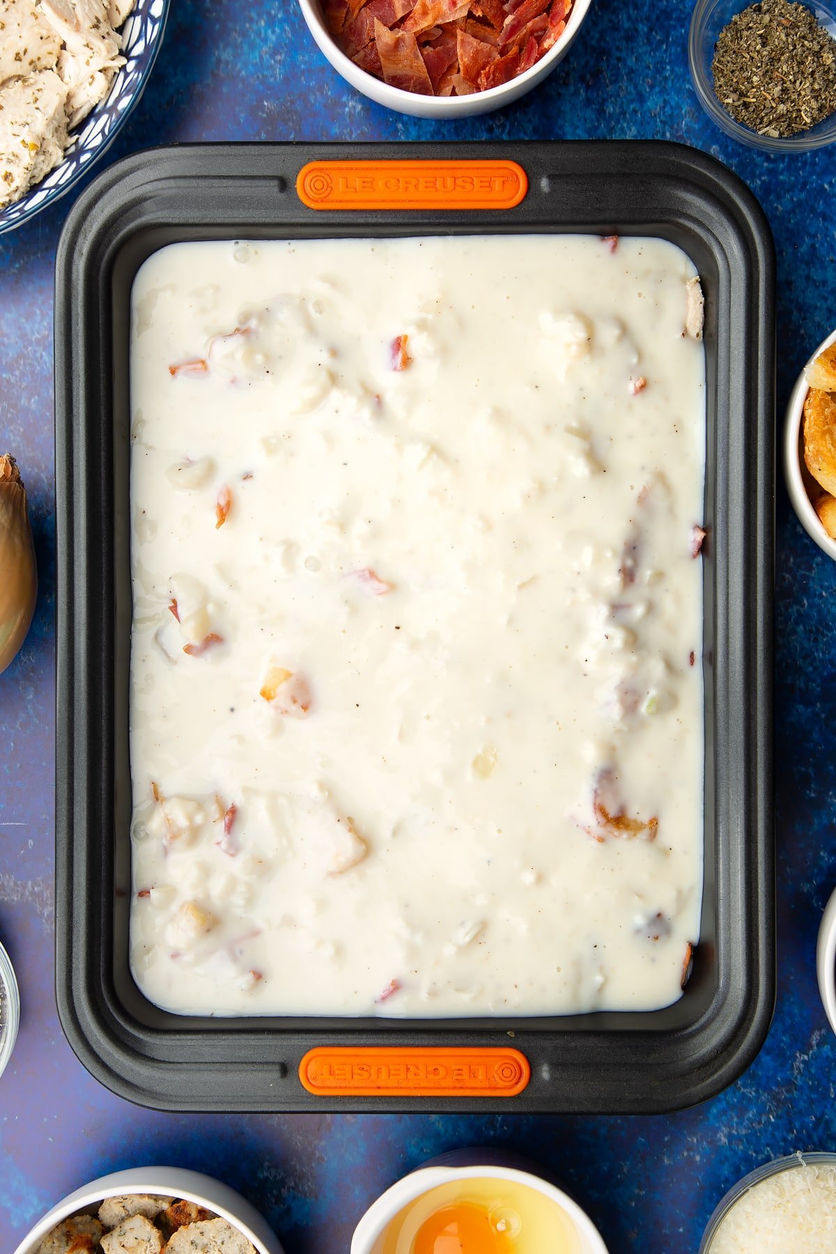 Chopped cooked turkey, stuffing, bacon and roast potatoes covered with white sauce in a roasting dish. Ingredients to make a leftover Christmas dinner pie surround the tray.