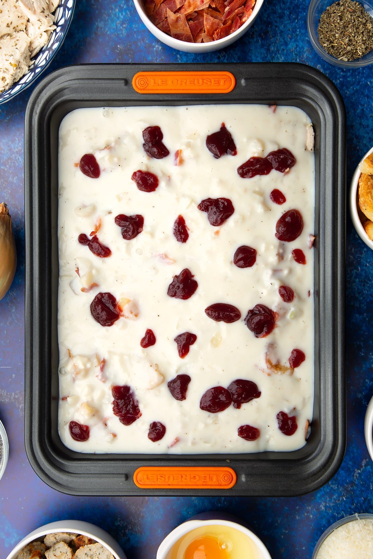 Chopped cooked turkey, stuffing, bacon and roast potatoes covered with white sauce and cranberry sauce in a roasting dish. Ingredients to make a leftover Christmas dinner pie surround the tray.