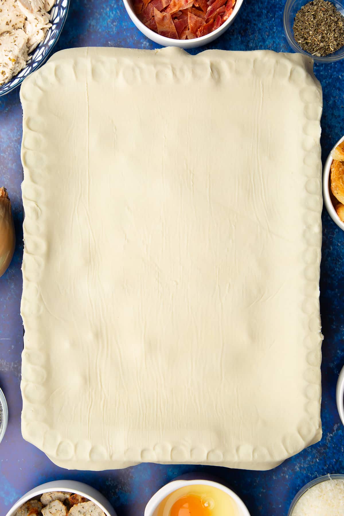 A roasting dish topped with a sheet of puff pastry. The edges have been crimped. Ingredients to make a leftover Christmas dinner pie surround the tray.