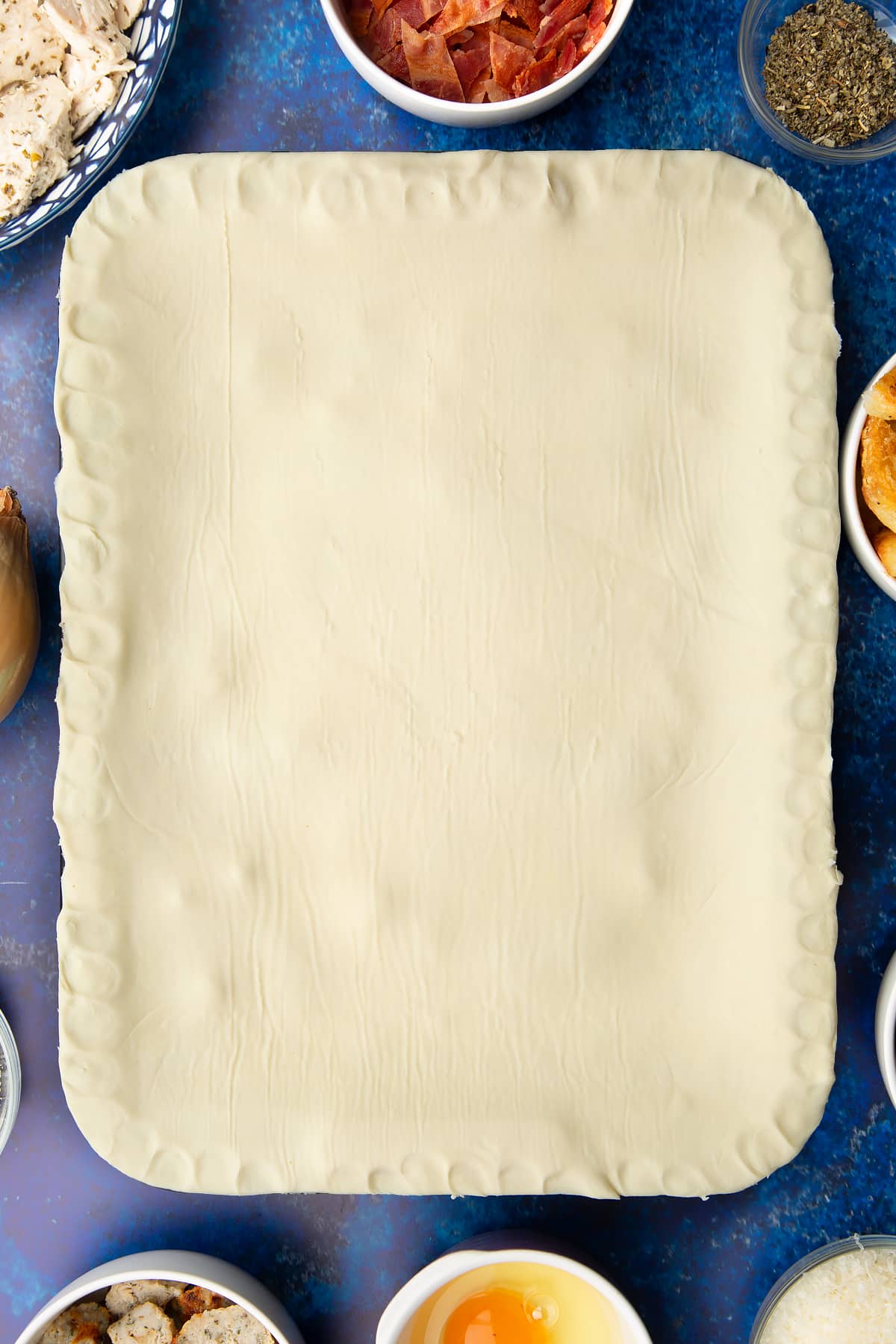 A roasting dish topped with a raw puff pastry lid. The edges have been crimped and trimmed. Ingredients to make a leftover Christmas dinner pie surround the tray.