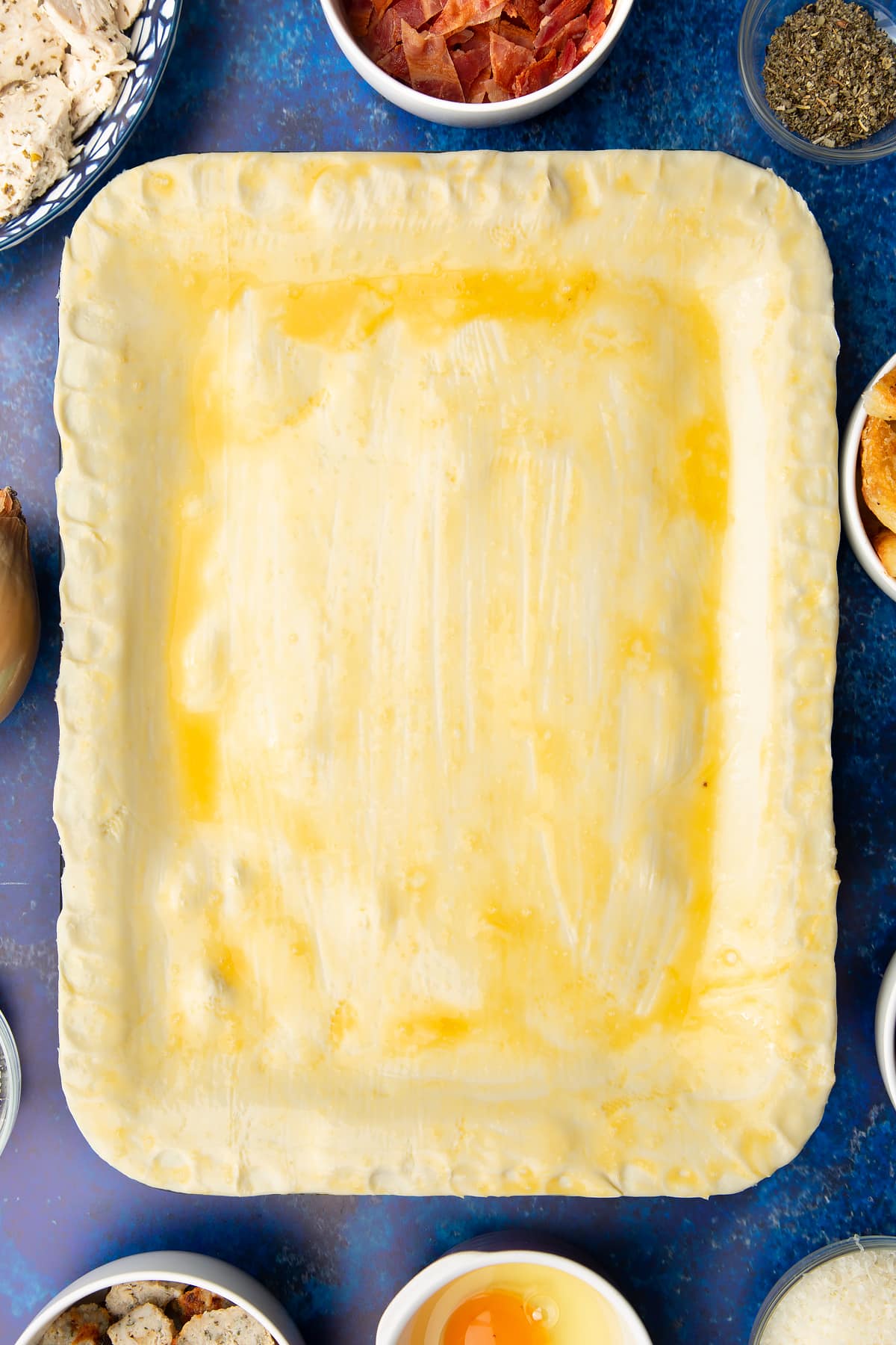 A roasting dish topped with a raw puff pastry lid that has been brushed with egg. Ingredients to make a leftover Christmas dinner pie surround the tray.