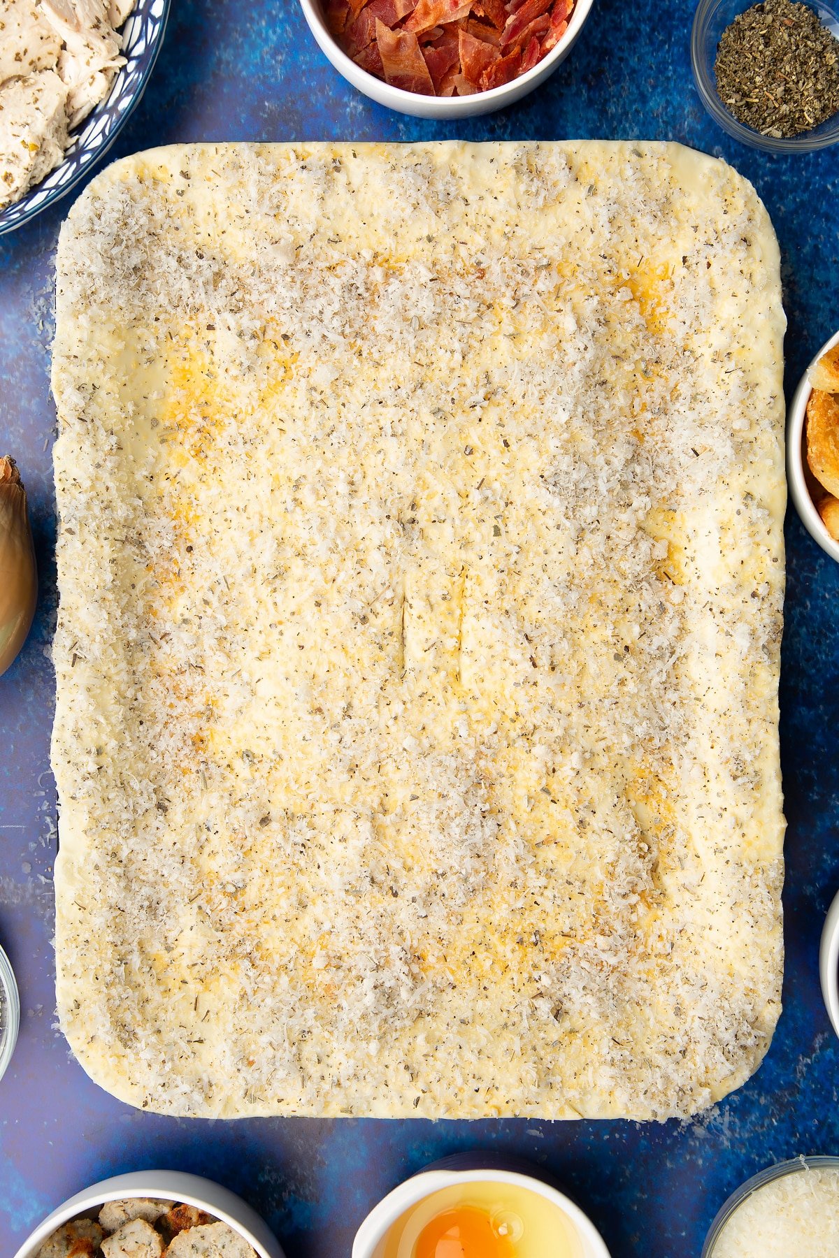 A roasting dish topped with a raw puff pastry lid that has been brushed with egg and sprinkled with breadcrumbs, cheese and herbs. Two slits have been cut in the centre.