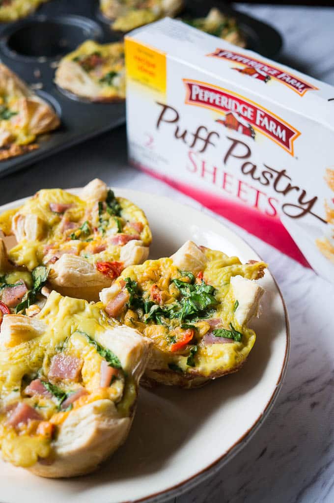 Three ham and cheese mini quiches made from puff pastry sitting on a plate, on a marble table top. The puff pastry sheets and the baking tin with more quiche cups sits in the background.