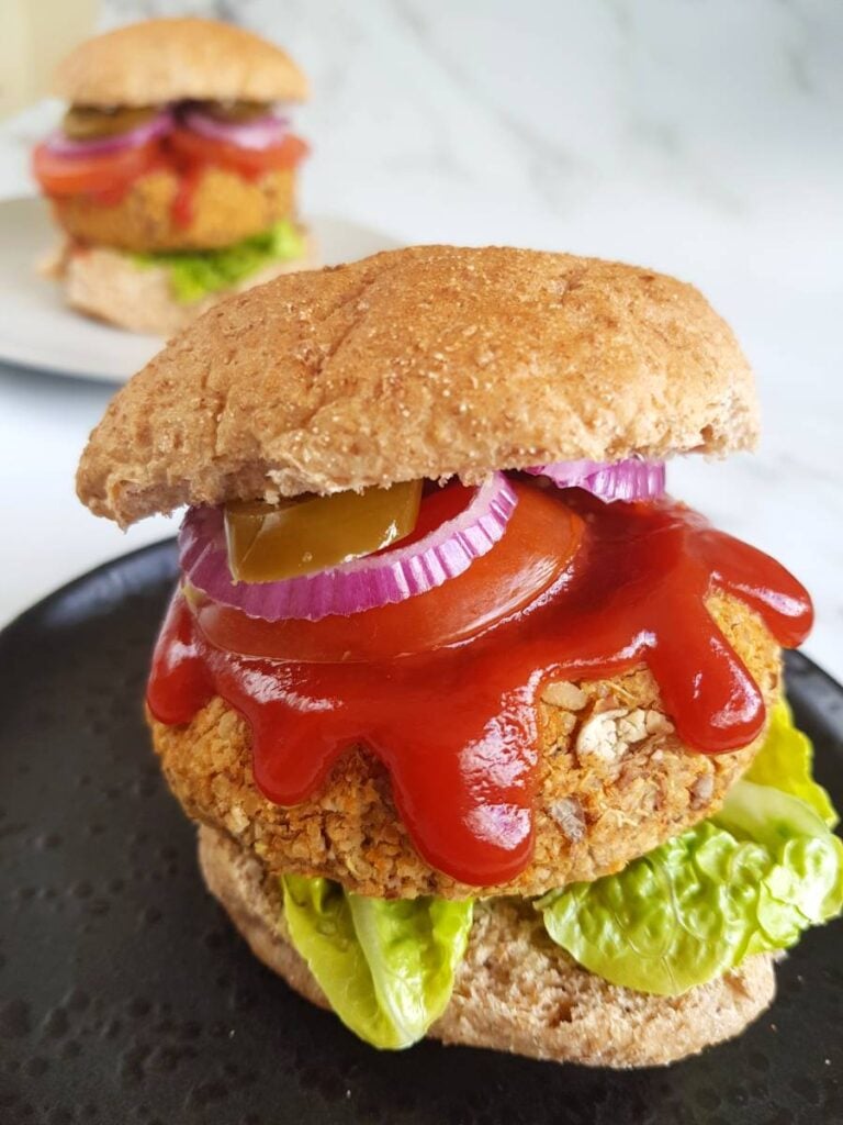 Pinto Bean Burgers with onions, tomatoes and ketchup on show. The burger is sat on a black plate on a white surface. In the background you can see another burger on a white plate.