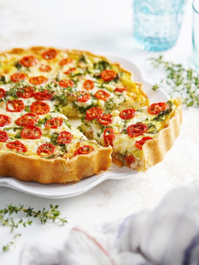 A slice of Shrimp Quiche (De Camarones) has gently been pulled away from the remainder of the quiche. This entire dish sits on a decorative white plate on a white background. At the side there's some leaves and two drinks glasses.