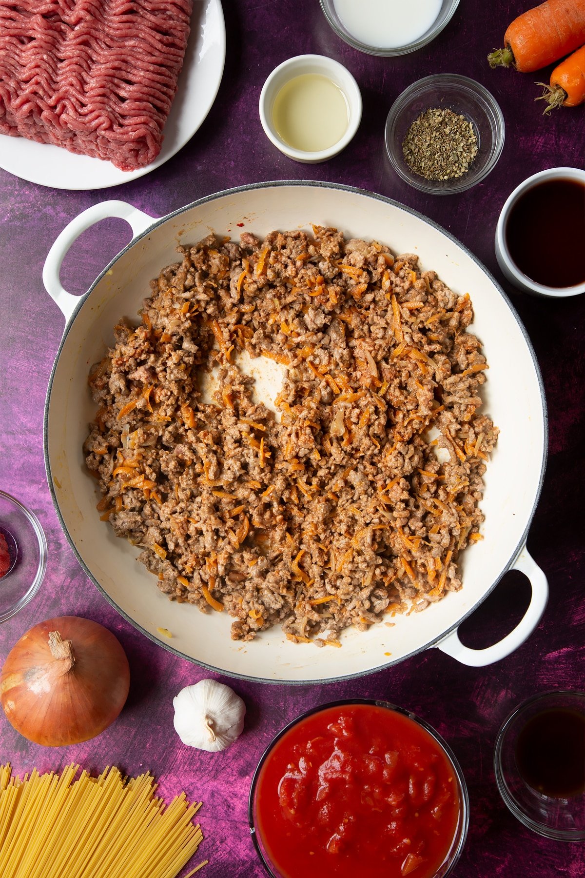 A large frying pan with fried carrot, onion, garlic, dried oregano, lean beef mince and tomato puree. Ingredients to make spaghetti bolognese Gordon Ramsay style surround the pan.