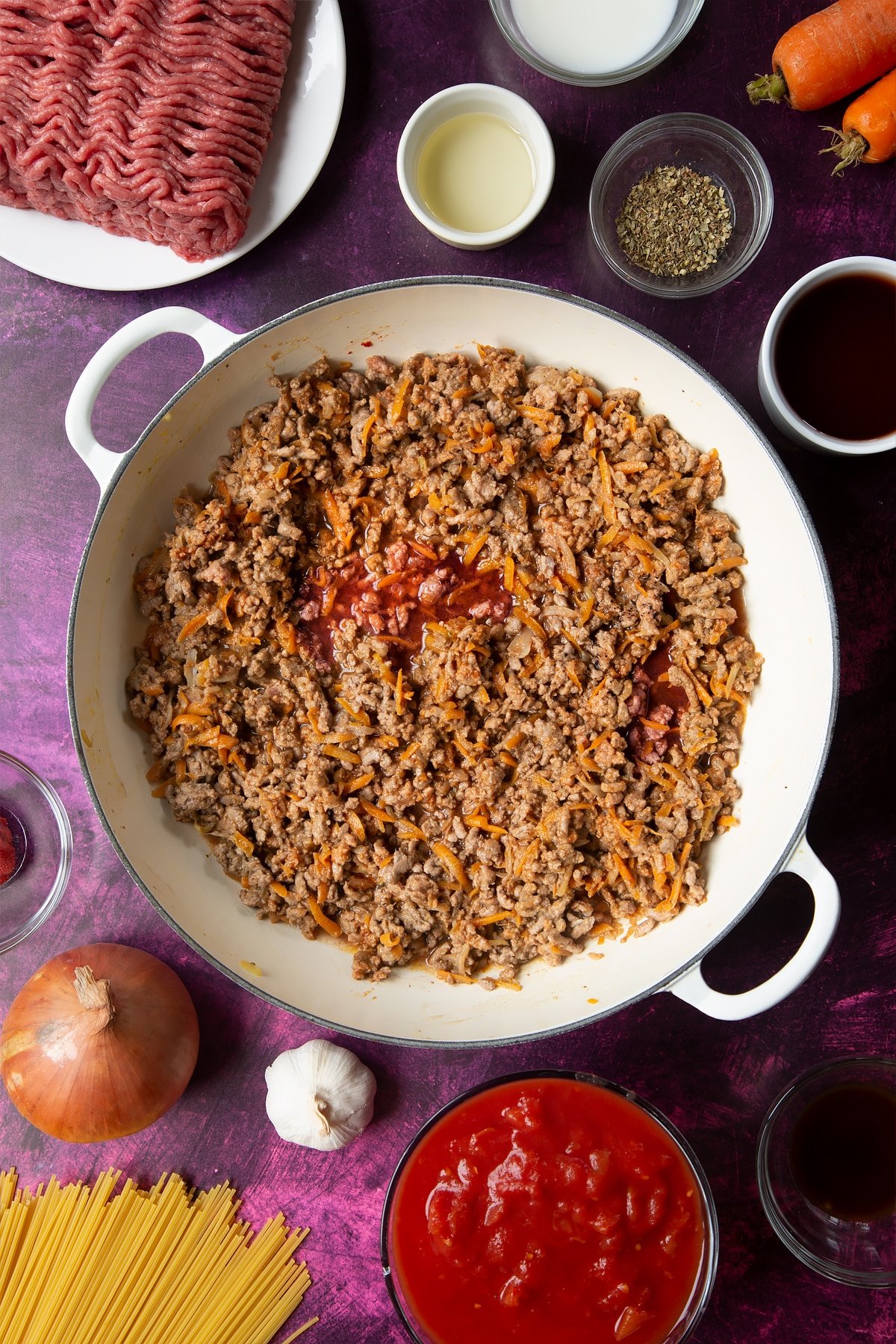 A large frying pan with fried carrot, onion, garlic, dried oregano, lean beef mince, tomato puree and red wine. Ingredients to make spaghetti bolognese Gordon Ramsay style surround the pan.