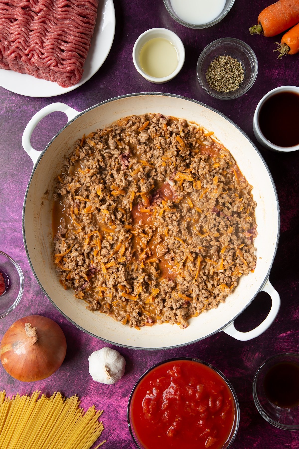 A large frying pan with fried carrot, onion, garlic, dried oregano, lean beef mince, tomato puree and reduced red wine. Ingredients to make spaghetti bolognese Gordon Ramsay style surround the pan.