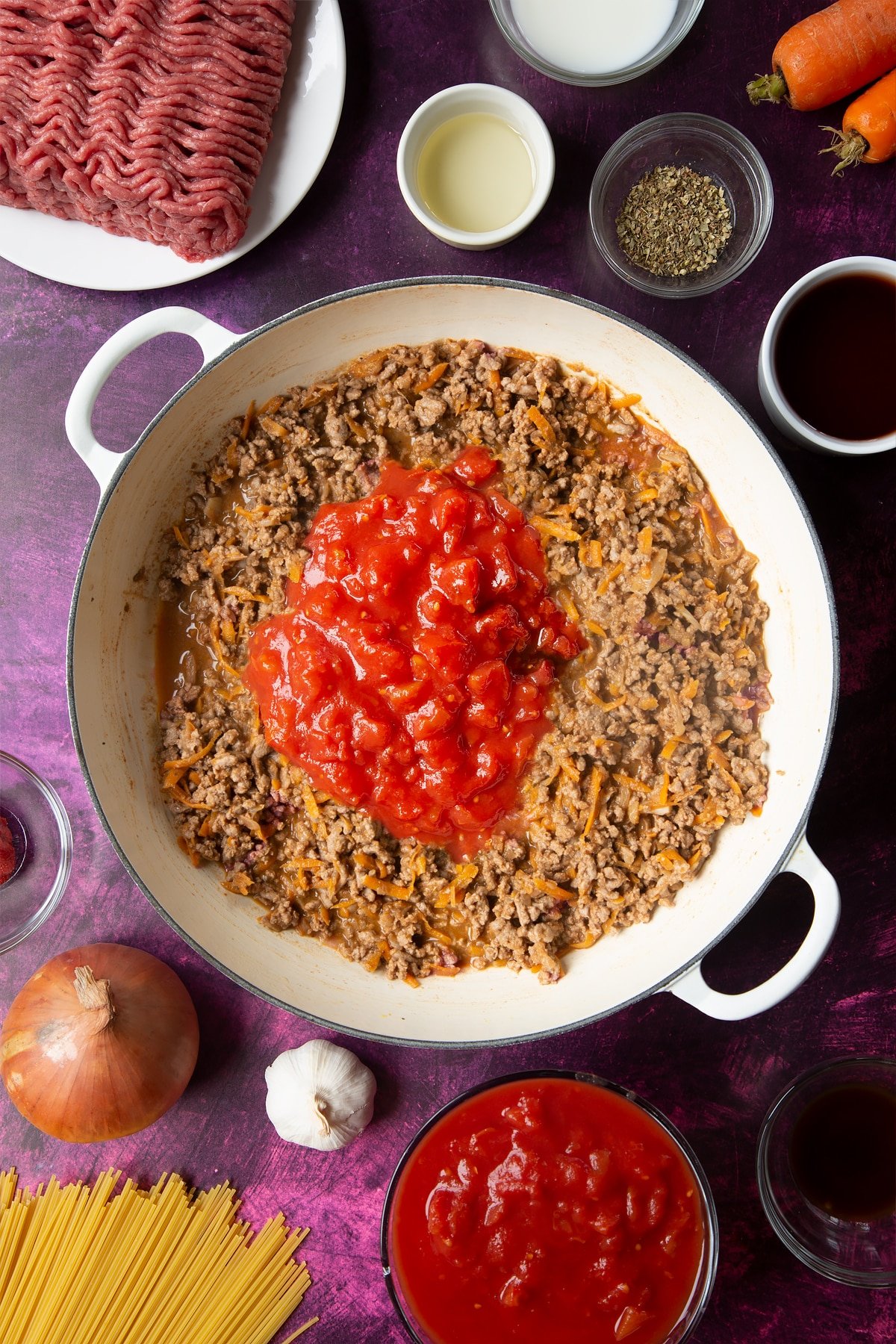 A large frying pan with fried carrot, onion, garlic, dried oregano, lean beef mince, tomato puree and red wine. Tinned tomatoes are added on top. Ingredients to make spaghetti bolognese Gordon Ramsay style surround the pan.