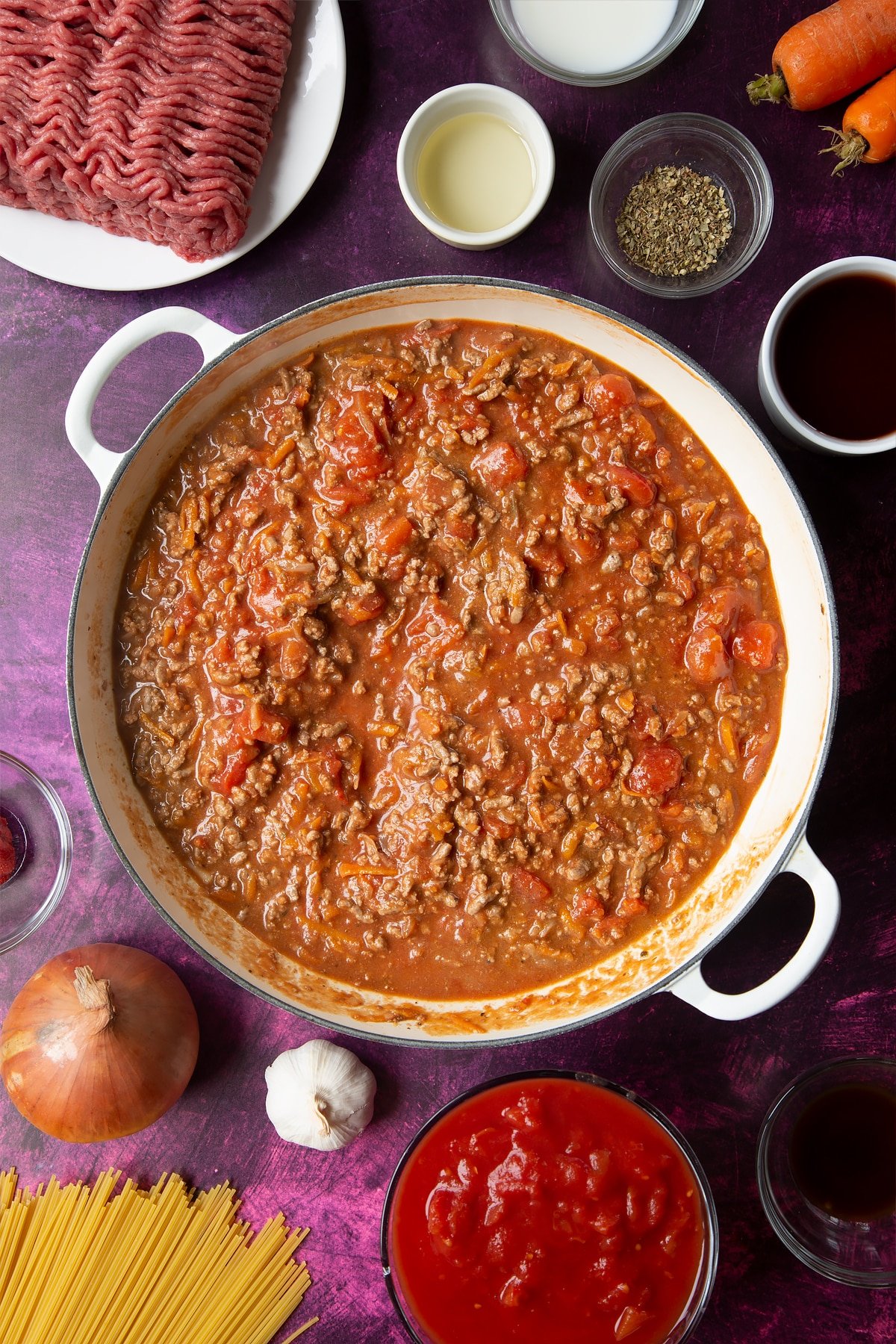 A large frying pan with fried carrot, onion, garlic, dried oregano, lean beef mince, tomato puree, red wine and tinned tomatoes. Ingredients to make spaghetti bolognese Gordon Ramsay style surround the pan.