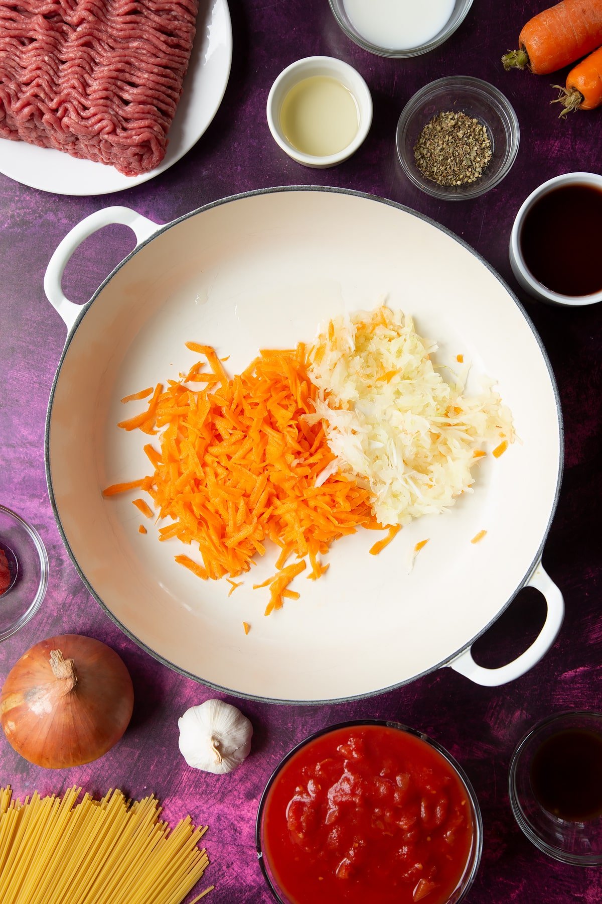 A large frying pan with olive oil, grated carrot and grated onion. Ingredients to make spaghetti bolognese Gordon Ramsay style surround the pan.